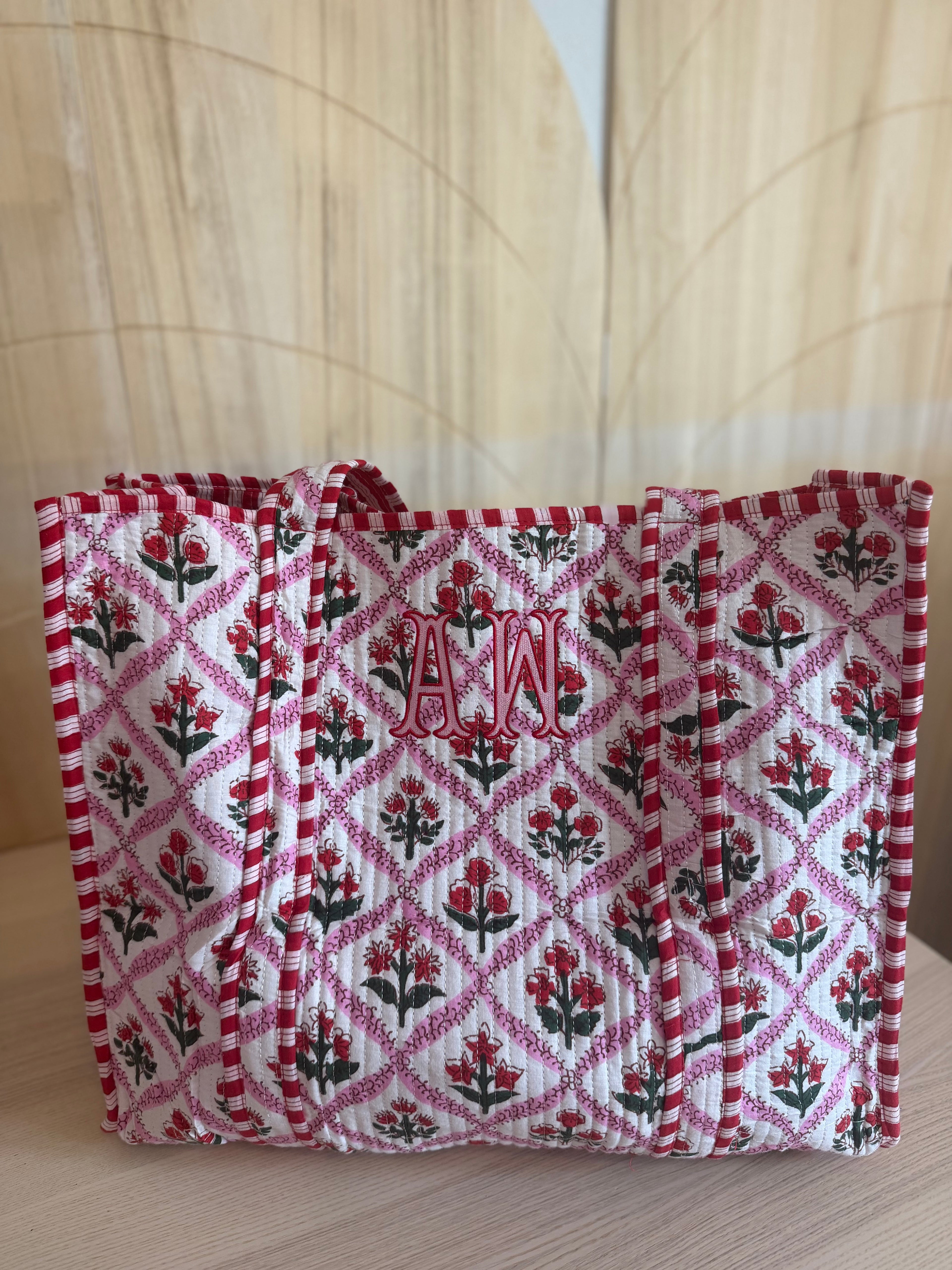 A quilted fabric tote bag with a red and white diamond lattice pattern, decorated with red and black floral motifs and personalized with a red monogram 'AW' on the front, set against a light wood background.