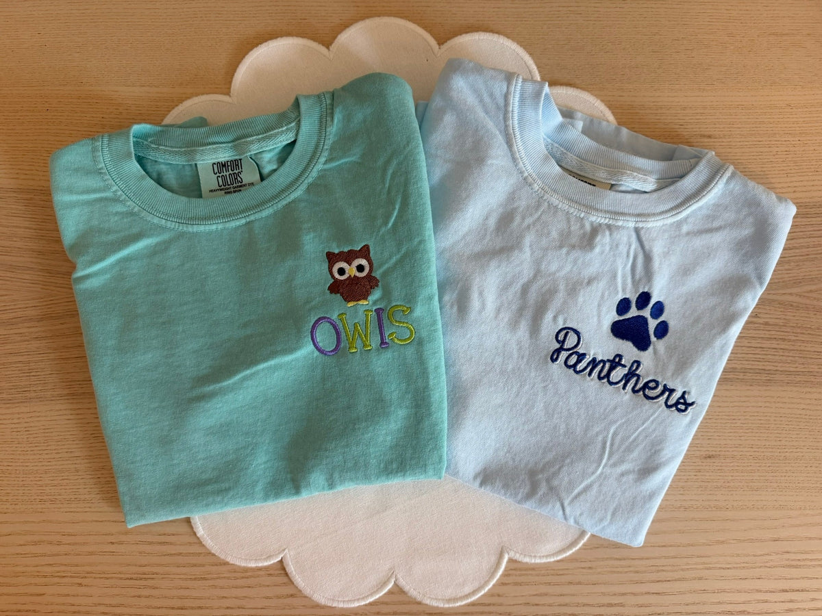 Two folded short-sleeve baby t-shirts on a scalloped white plate over a wooden surface. The top shirt is mint green with an owl graphic and the text 'OWLS'.
